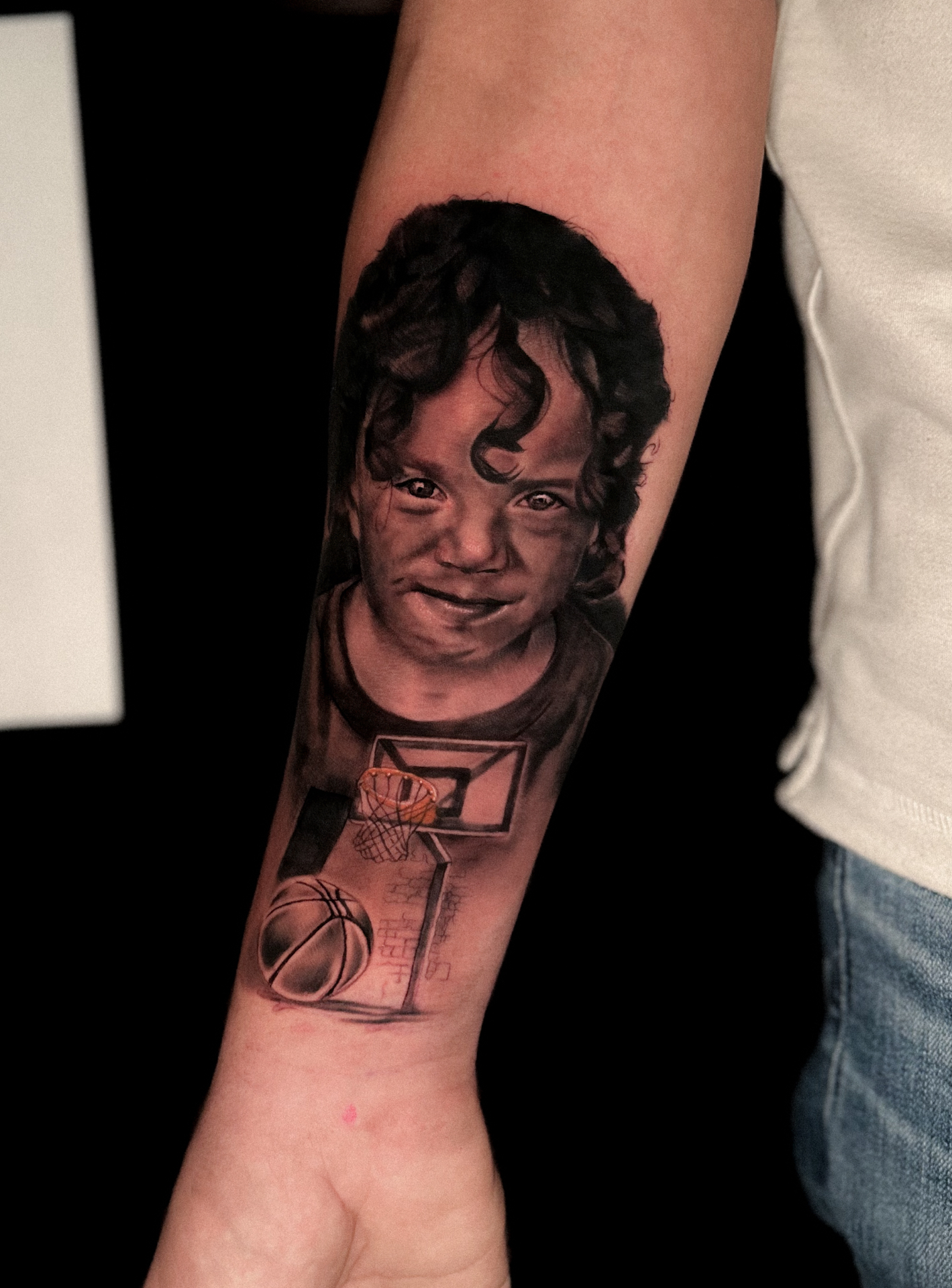 Black and grey child portrait forearm tattoo with a basketball hoop by Ramón Santiago at R INKS ARTS