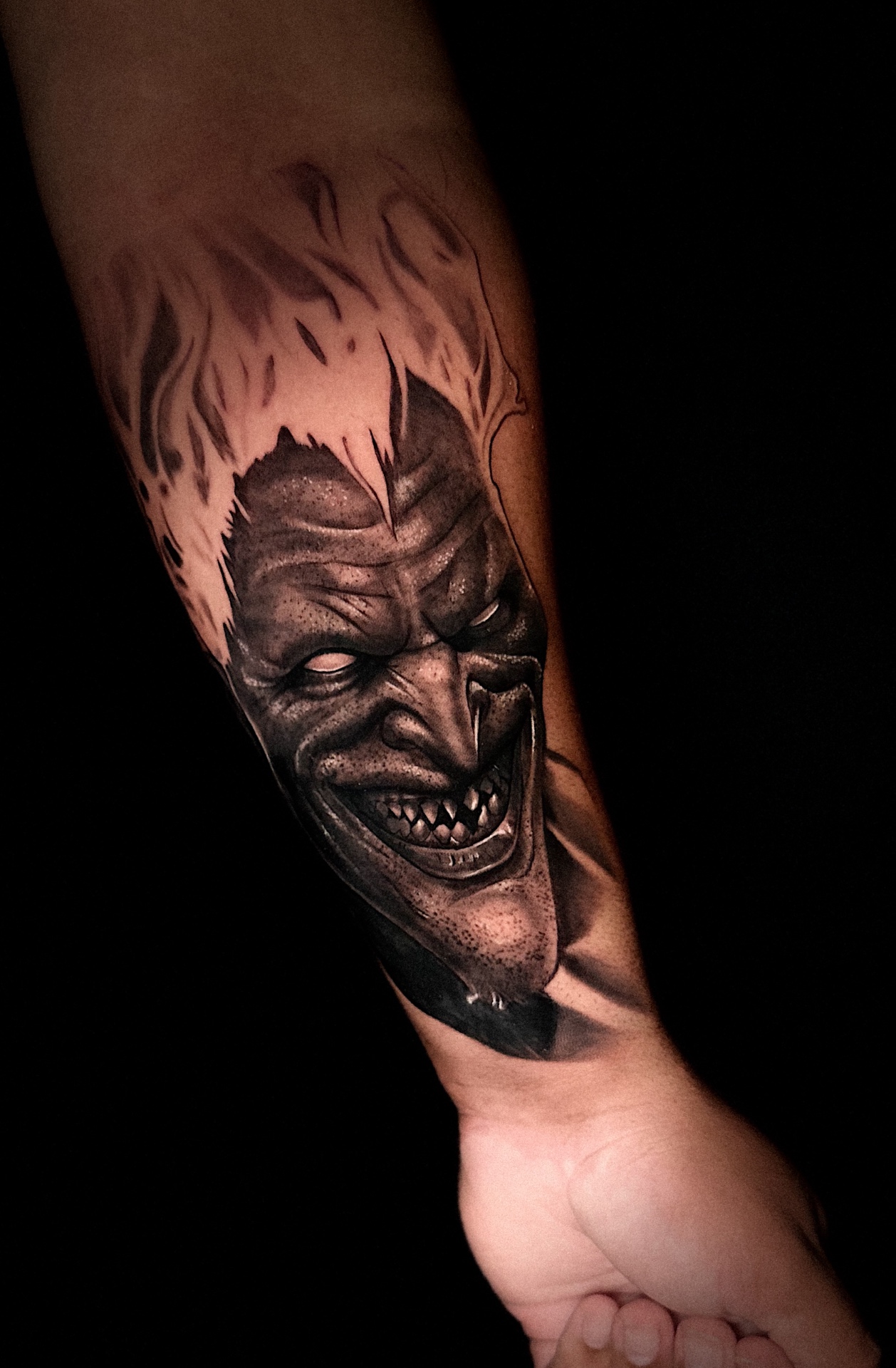 Black and grey grinning joker forearm tattoo by Ramón Santiago at R INKS ARTS