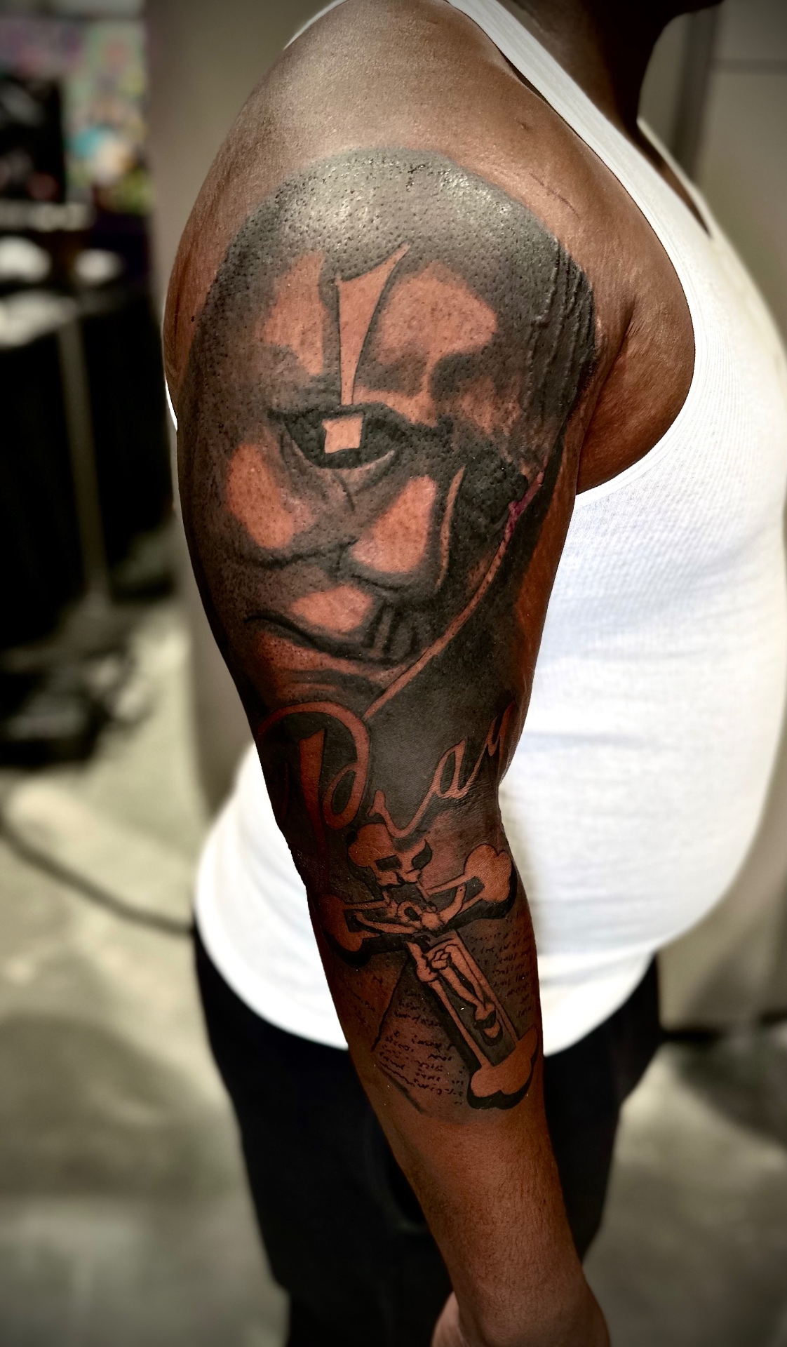 Black and grey memorial shoulder tattoo with a face, cross and script lettering by Ramón Santiago at R INKS ARTS